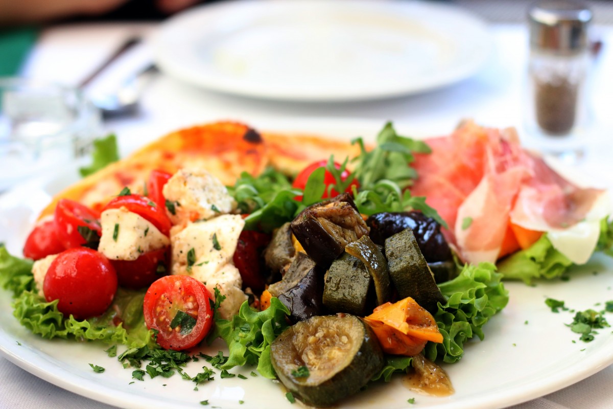 Plate with salad of cherry tomatoes and mozzarella, grilled vegetables, prosciutto, and lettuce on a white tablecloth.